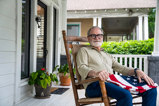 Retired veteran looking at camera while sitting on his rocking chair on his porch with a flag on his lap.